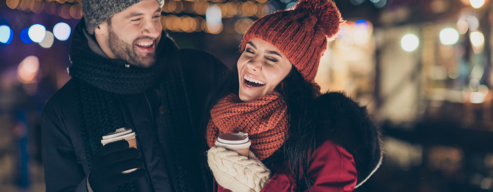 Photo of two people with hot beverage spending last evening in year, outdoors having best time wearing warm jackets knitted hats and scarfs